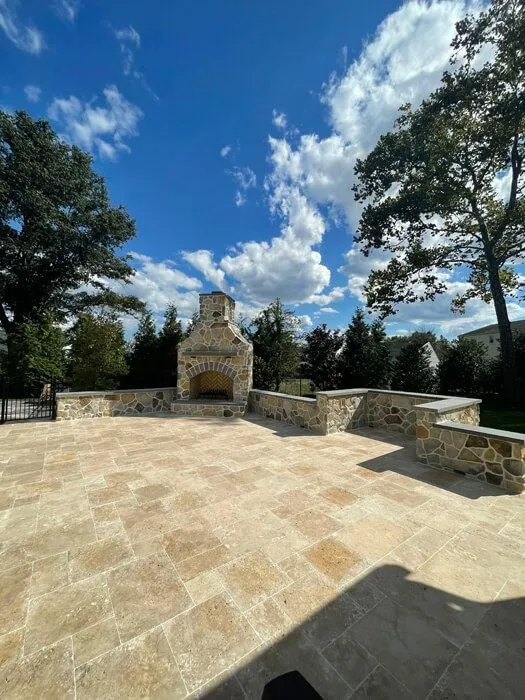Large tan paver patio with a stone fireplace and seat walls under a blue sky
