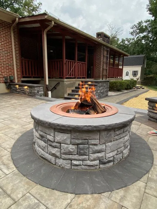 Round stone fire pit with a fire burning on a new paver patio in front of a brick ranch home