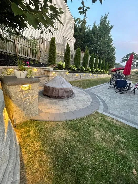 Lit stone seat wall and covered fire pit on a round paver patio at dusk